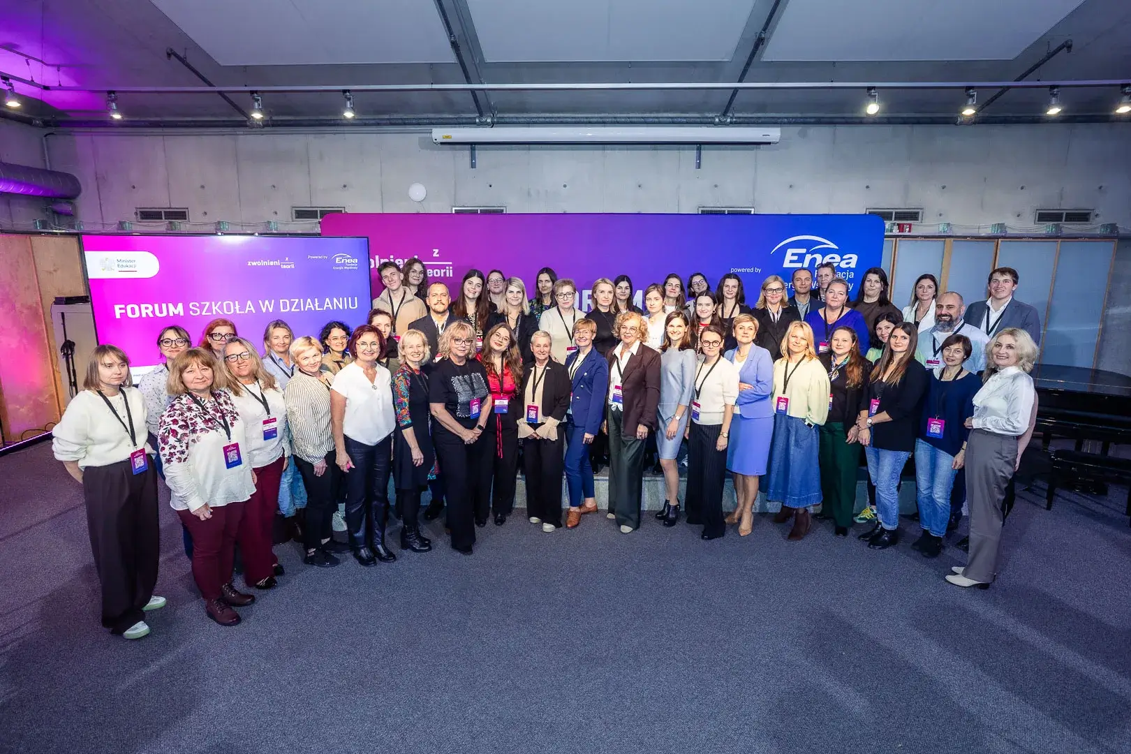 Grupowe zdjęcie uczestników i uczestniczek wydarzenia Forum Szkoła w Działaniu. Kilkadziesiąt osób stoi przed dużą, różowo-niebieską ścianką z logotypami Zwolnieni z Teorii, Fundacji Enea - Energia Wspólnoty i Minister Edukacji. Wszyscy patrzą w stronę aparatu i uśmiechają się. Zdjęcie wykonane w jasnym, industrialnym wnętrzu z widocznym oświetleniem sufitowym.
