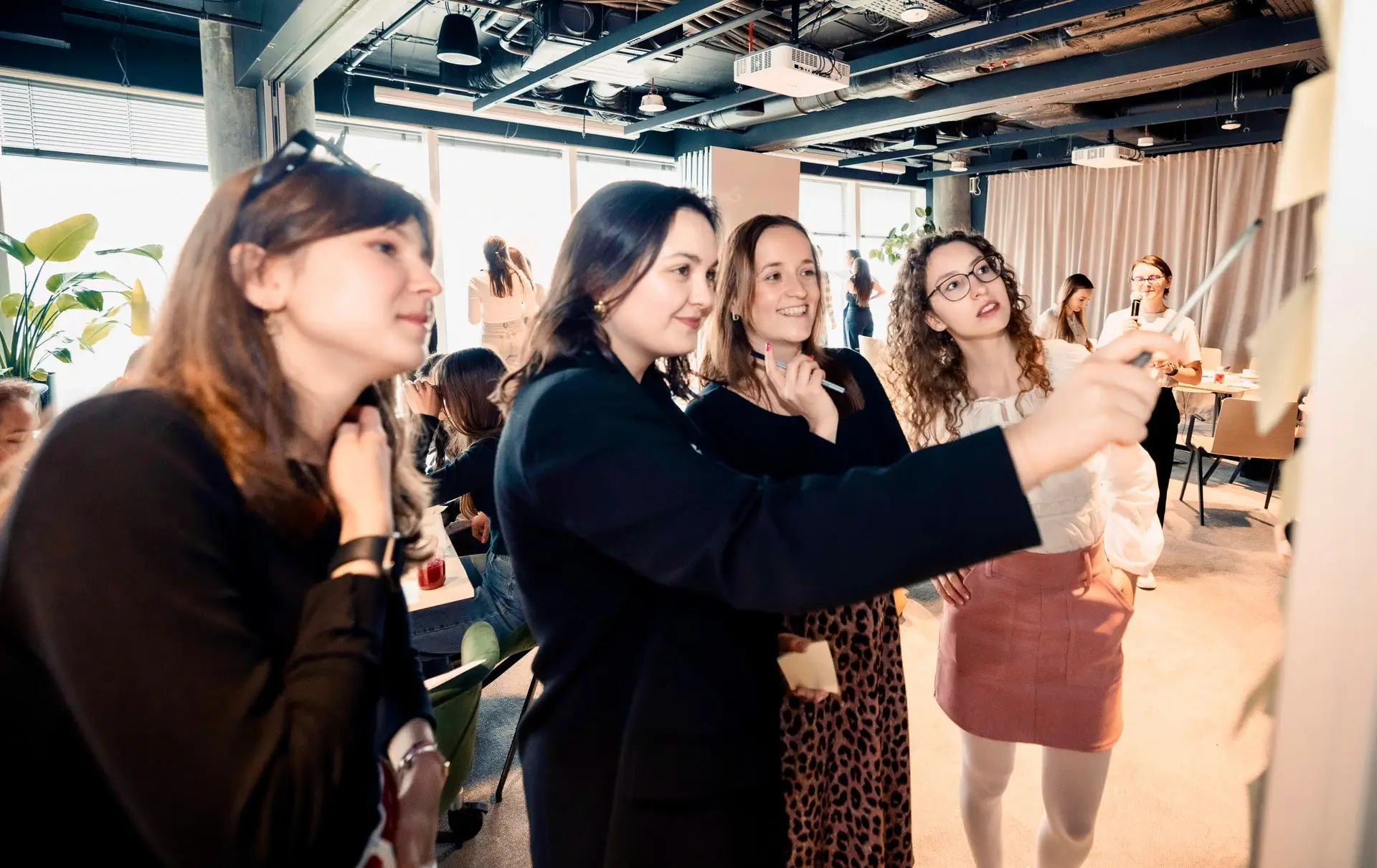 Grupa młodych osób stojących razem i patrzących w stronę tablicy z karteczkami samoprzylepnymi w jasnym biurowym wnętrzu.