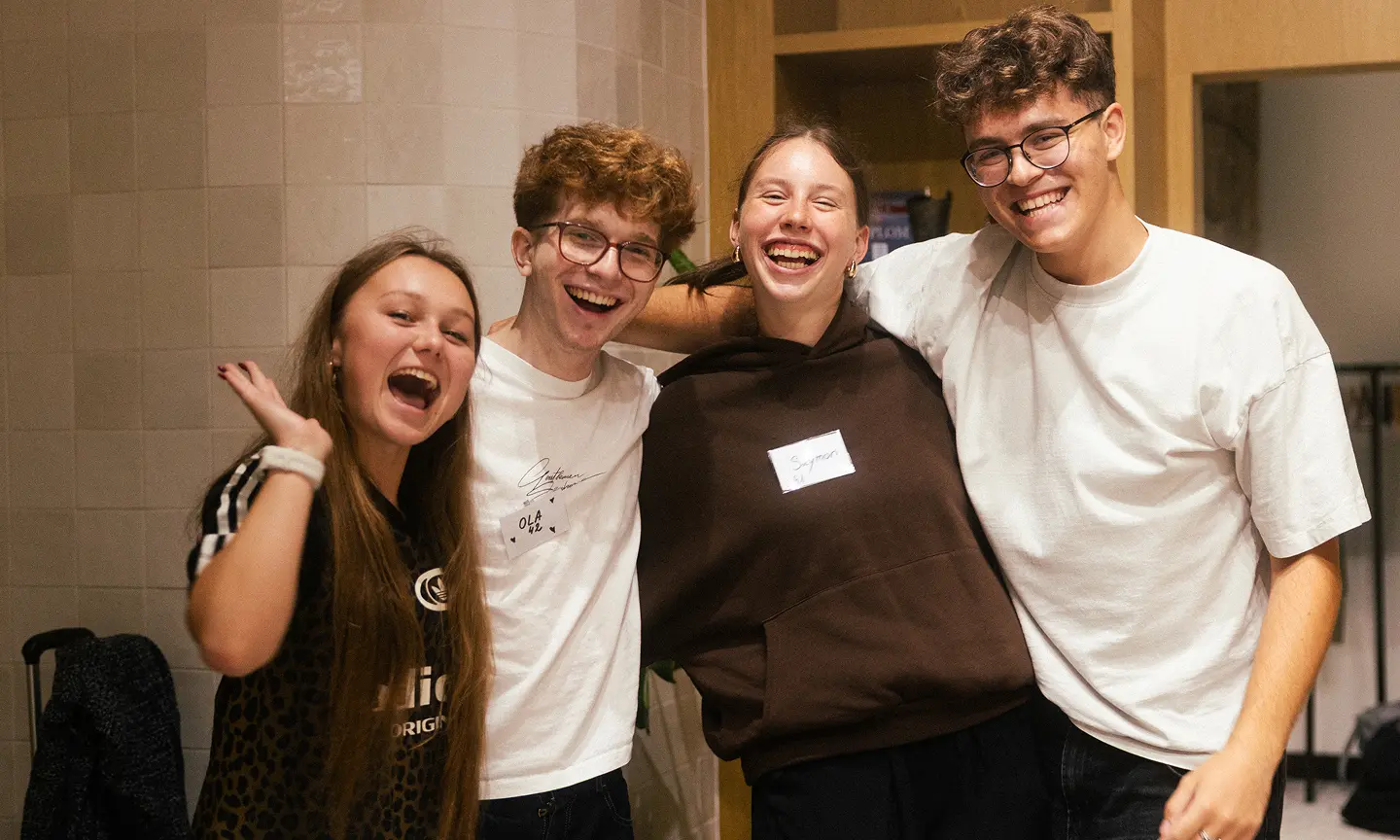 Zdjęcie grupy 4 młodych ludzi uśmiechających się i obejmujących się nawzajem w ciepłej, przytulnej przestrzeni.