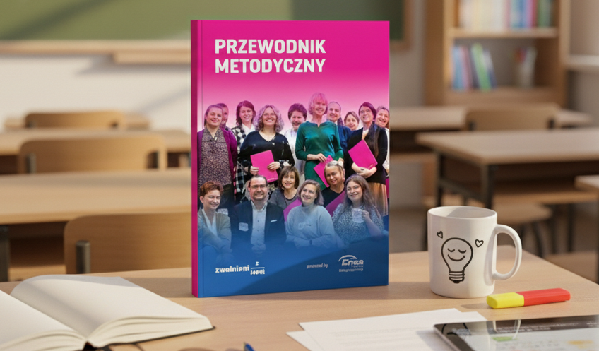 Materiał zatytułowany&nbsp;Przewodnik metodyczny&nbsp;stojący na biurku w szkolnej klasie, obok kubka z uśmiechniętą buźką w kształcie żarówki oraz otwartych zeszytów, karteczek oraz tabletu. W tle widoczne są ławki szkolne oraz tablica kredowa.
