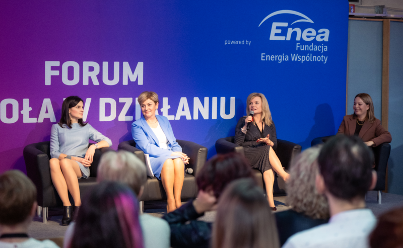 Cztery kobiety siedzą na scenie podczas panelu dyskusyjnego Forum Szkoła w Działaniu; jedna z nich trzyma mikrofon i mówi, a publiczność słucha. W tle widoczne jest fioletowo-niebieskie tło z logo Fundacji Enea - Energia Wspólnoty.