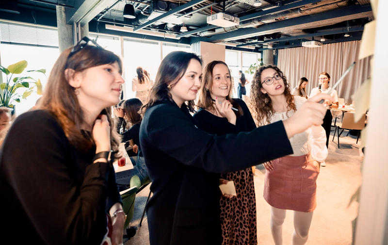 Cztery kobiety stojące wokół tablicy i wspólnie pracujące nad zadaniem podczas warsztatu; jedna z nich zapisuje wskazuje długopisem na zapisane karteczkach samoprzylepne. W tle widać także innych uczestników warsztatu.